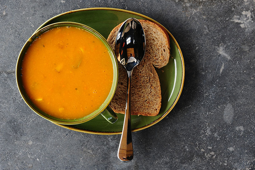 Pompoensoep in een schaaltje op een bord met een lepel en een sneetje brood